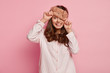 © wayhome.studio  - Pleased delighted woman looks through warm eyemask, dressed in nightclothes, poses against pink background, looks with one eyes, shows white teeth. People, bedtime, rest and positiveness concept