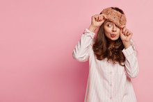 Indoor Shot Of Funny Young European Woman Wears Eyemask, Pyjamas, Looks Away, Poses Against Pink Background With Copy Space For Your Promotional Content Or Advertisement. People And Rest Concept