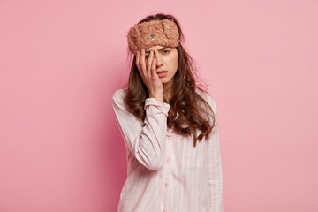 Upset tired young woman has not enough sleep, touches face with hand, wears eyemask and casual jumper, isolated over pink background, slept bad at night. People, tiredness and awakening concept