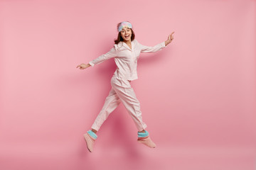 Wall Mural - Isolated shot of joyful young woman with happy expression, jumps in air, dressed in pyjamas and domestic slippers, spreads hands, poses over pink background, smiles broadly. Good morning concept