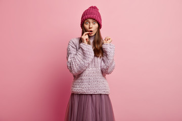 Wall Mural - Puzzled scared hipster girl has bugged eyes, keeps fore finger on lips, wears winter hat, oversized long sleeved jumper, skirt, models over rosy background, has overwhelmed facial expression