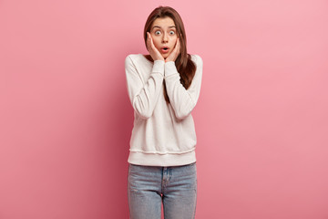 Wall Mural - Photo of emotive beautiful young woman touches cheeks with both palms, dressed in casual outfit, reacts on something shocking, has eyes popped out, isolated on pink background, recieves bad news