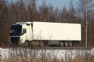 European white semi-trailer truck with logo copy space for writing on winter road on forest trees and blue sky background - side front view