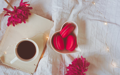 Top view of a heart shaped bowl and red macarons, light chain, a cup of black coffee on a book and two pink flowers on a bed or white textile background. Good morning coffee on Valentine's day concept