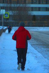 A man in a red jacket is jogging along a winter city embankment on a frosty winter day.