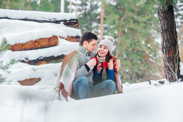 Wall Mural - Smiling young couple with cups sitting in snow park. Drinking hot drinks, having fun.