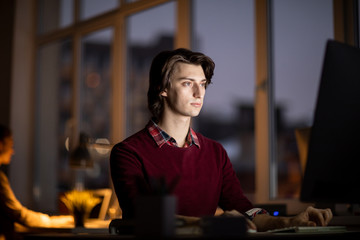 Wall Mural - young serious businessman in casualwear looking at computer screen while sitting by workplace in off