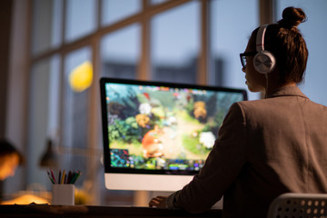 Young female colorist or footage editor in headphones sitting in front of computer monitor with video on screen