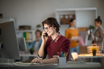 Poster - Smiling young man in casualwear sitting in front of computer monitor consulting with his colleague by smartphone