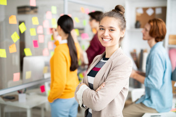Poster - Prety young cheerful office worker with crossed arms looking at you in working environment among colleagues
