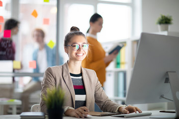 Happy casual businesswoman looking at you while sitting by desk in front of computer screen in working environment