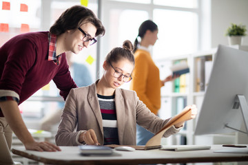 Fototapete - Young businesswoman and her colleague reading working points in notepad while organizing work