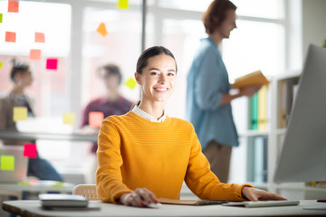 Poster - Pretty cheerful businesswoman in casualwear sitting by desk in front of computer monitor and carrying out her work