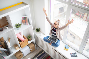 Poster - Ecstatic businesswoman with laptop raising her arms while sitting on windowsill and expressing victory