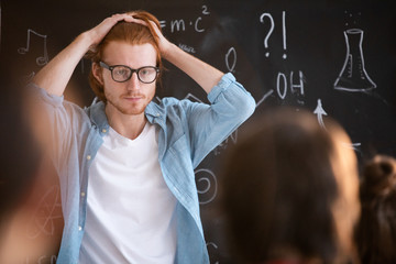 Fototapete - Young confused man in casualwear touching his head while standing by blackboard with written formula and looking at audience