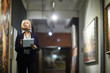 Portrait of mature woman holding clipboard looking at paintings standing in art gallery or museum, copy space