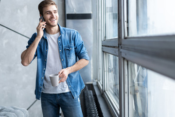 Smiling young man talking on mobile phone, looking at the window at home.