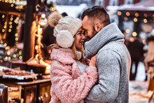An Attractive Couple In Love, A Stylish Couple Wearing Warm Clothes Cuddling Together And Looking Each Other At The Winter Fair At A Christmas Time.