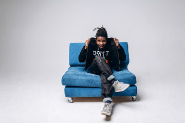 Wall Mural - One attractive smiling black man in a black jacket and black pants on a gray background sidin on a blue sofa