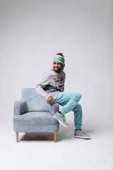 Wall Mural - Portrait of a smiling black young model in a gray sweater and blue pants on a gray background near the chair