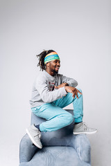 Wall Mural - Portrait of a smiling black young model in a gray sweater and blue pants on a gray background near the chair