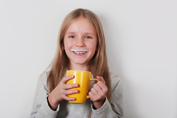 Cute happy blond little girl drinking milk or yogurt from a yellow cup on a white background. Mustache from yogurt or milk. Dairy products concept