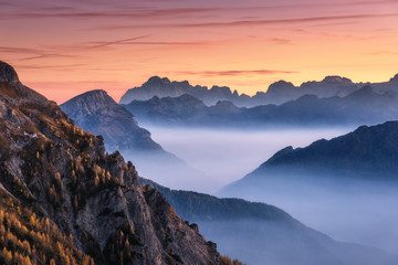 Wall Mural - Mountains in fog at beautiful sunset in autumn in Dolomites, Italy. Landscape with alpine mountain valley, low clouds, trees on hills, orange sky with clouds at dusk. Aerial view. Passo Giau. Nature
