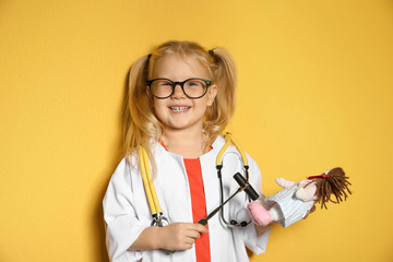 Wall Mural - Cute child imagining herself as doctor while playing with reflex hammer and doll on color background