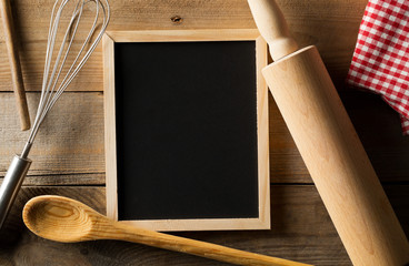 Wall Mural - Blank, empty, black chalkboard with wooden rolling pin, wire whisk and red checkered dish towel flat lay from above on brown wooden table