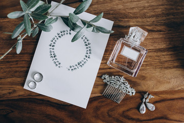 on wooden table are the wedding rings of the bride and groom, which lie on the invitation card with a pattern, next to it is a sprig of eucalyptus, a bottle of perfume and earrings made of white gold