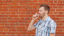 Fashionable And Stylish Guy With A Beard Smoking A Vape Against A Brick Wall, Copy Space, Slow-mo, Outside