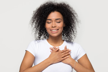 Grateful hopeful happy black woman holding hands on chest feeling pleased thankful, sincere african lady expressing heartfelt love appreciation gratitude honesty isolated on white studio background