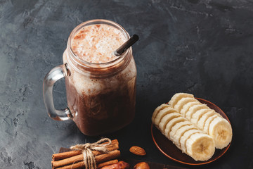 Chocolate smoothie in a jar, protein milkshake, nuts, banana slices, dates, cinnamon sticks, healthy eating on a dark background