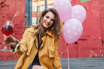 Young beautiful smiling woman in orange trench coat holding cocktail in hand happily looking in camera with pink balloons near in old courtyard of cafe