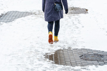 Wall Mural - Winter Walk in Yellow Leather Boots. Back view on the feet of a women walking along the icy snowy pavement. Pair of shoe on icy road in winter. Abstract empty blank winter weather background