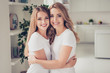 Close up photo cheer toothy smiling two people mum and teen daughter holding hands arms around chest lovely looking best buddies rejoice wear white t-shirts jeans in bright kitchen