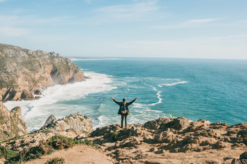 Wall Mural - A tourist with a backpack on a cliff against the background of the Atlantic Ocean raised his hands and shows how he is free and happy.