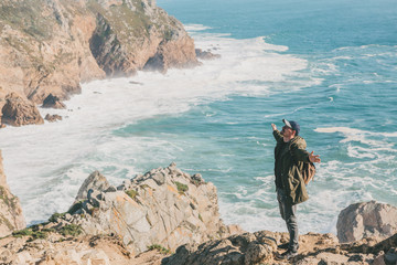 Wall Mural - A tourist with a backpack on a cliff against the background of the Atlantic Ocean raised his hands and shows how he is free and happy.