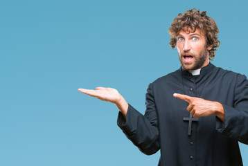 Poster - Handsome hispanic catholic priest man over isolated background amazed and smiling to the camera while presenting with hand and pointing with finger.