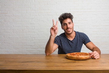 Wall Mural - Young handsome and natural man sitting on a table fun and happy, positive and natural, doing a gesture of victory, peace concept. Eating a delicious pizza.