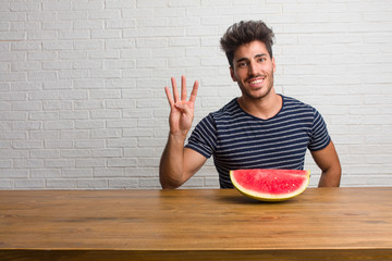 Young handsome and natural man sitting on a table showing the number four, symbol of counting, concept of mathematics, confident and cheerful. Eating a watermelon.