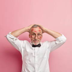 Wall Mural - Vertical shot of funny bearded senior male model keeps both hands on head, wears white shirt and round spectacles, poses over rosy background, has kiss marks on face, being in love with colleague