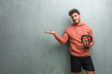Fototapeta Łazienka - Young fitness man against a grunge wall holding something with hands, showing a product, smiling and cheerful, offering an imaginary object. Holding an iron dumbbell.