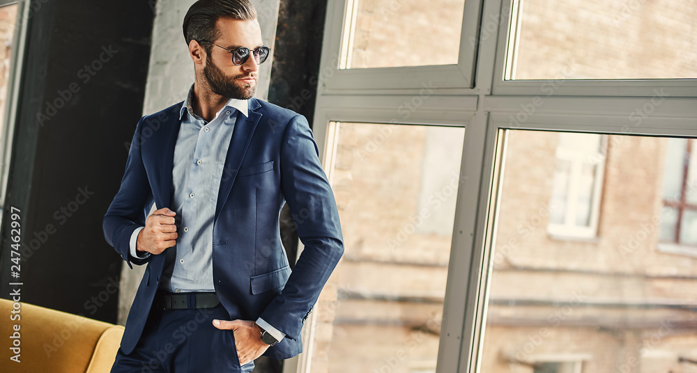 Confident and stylish. Businessman in sunglasses and fashion suit is thoughtfully looking in window while standing at office - obrazy, fototapety, plakaty 