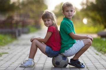 Two cute blond happy young children, smiling boy and long-haired girl sitting back-to-back on soccer ball on empty sunny suburb paved road blurred summer bright background. Children games concept.