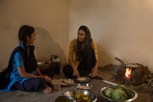 Rural Woman Sitting In Kitchen Cooking Food On Firewood With Utensils And Vegetables On The Floor And Talking To Her Daughter.	