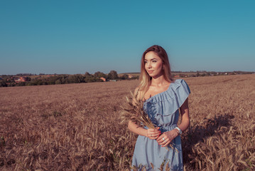 Girl in a wheat field in the summer in a blue dress and wheat in the hand of ears of corn. A woman walks across field, long hair casual makeup. Free space for text.