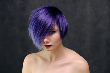 A beautiful, sexy girl with purple hair and a short haircut sits in the middle of the photo with a gray background