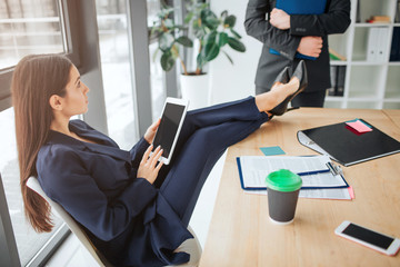 Young businesswoman sit on chair at window in room. She hold legs on table and look at guy. Young man stand in front of her. Model hold tablet and play.