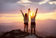 Happy Couple With Raised Arms Stands On Mountain Top Against Sunset And Having Fun In Winner Pose